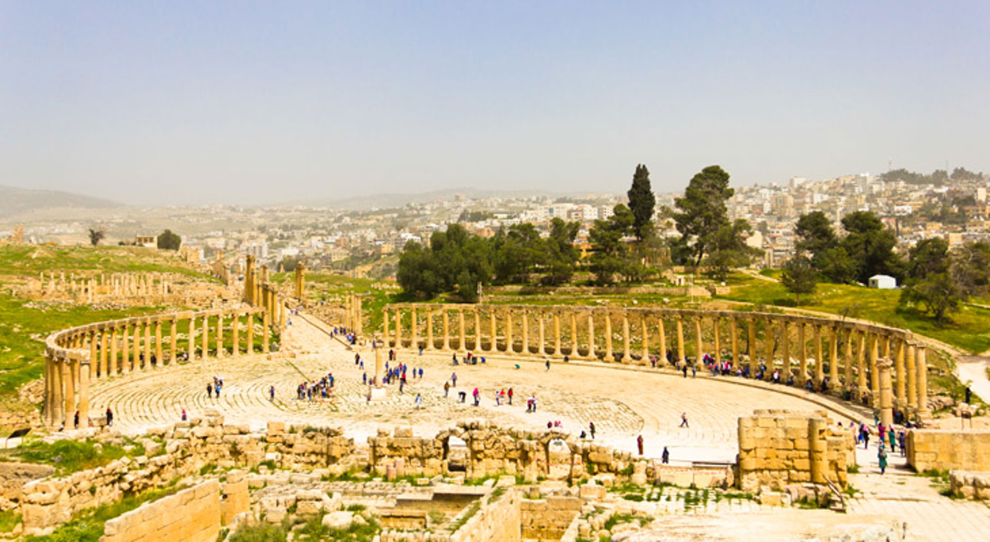 Romeins amfitheater met zuilengalerij en bezoekers, met uitzicht op de stad op de achtergrond.