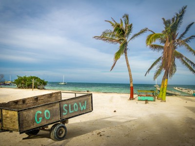 Palmbomen en wit zandstrand aan zee in Belize, met houten kar waarop “GO SLOW” staat.