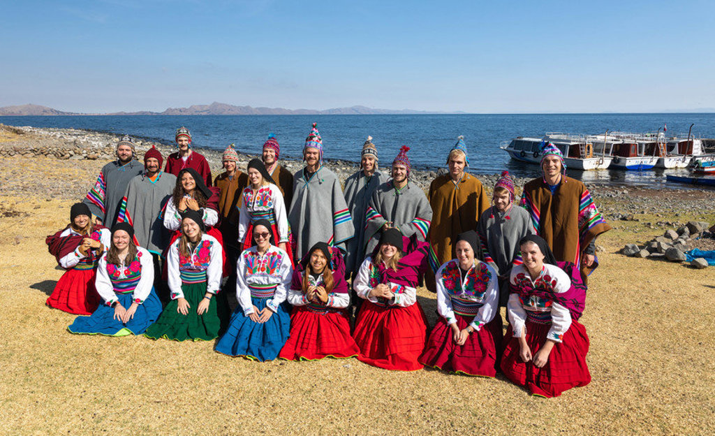 Groep 22-35ers in traditionele kleding aan het Titicacameer in Peru, met boten op de achtergrond.