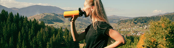 Vrouw drinkt uit herbruikbare waterfles tijdens wandeling met uitzicht op bergen en bos.