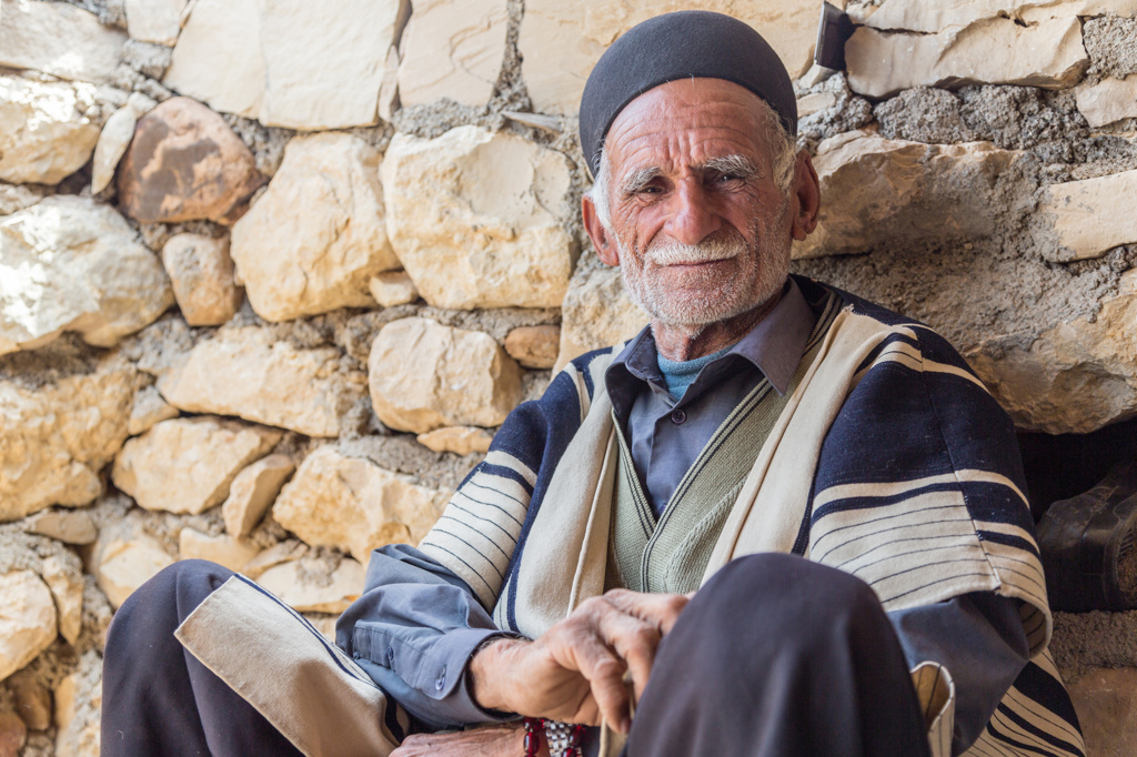 Portret van een oudere man met traditionele muts, zittend tegen een stenen muur in Iran.