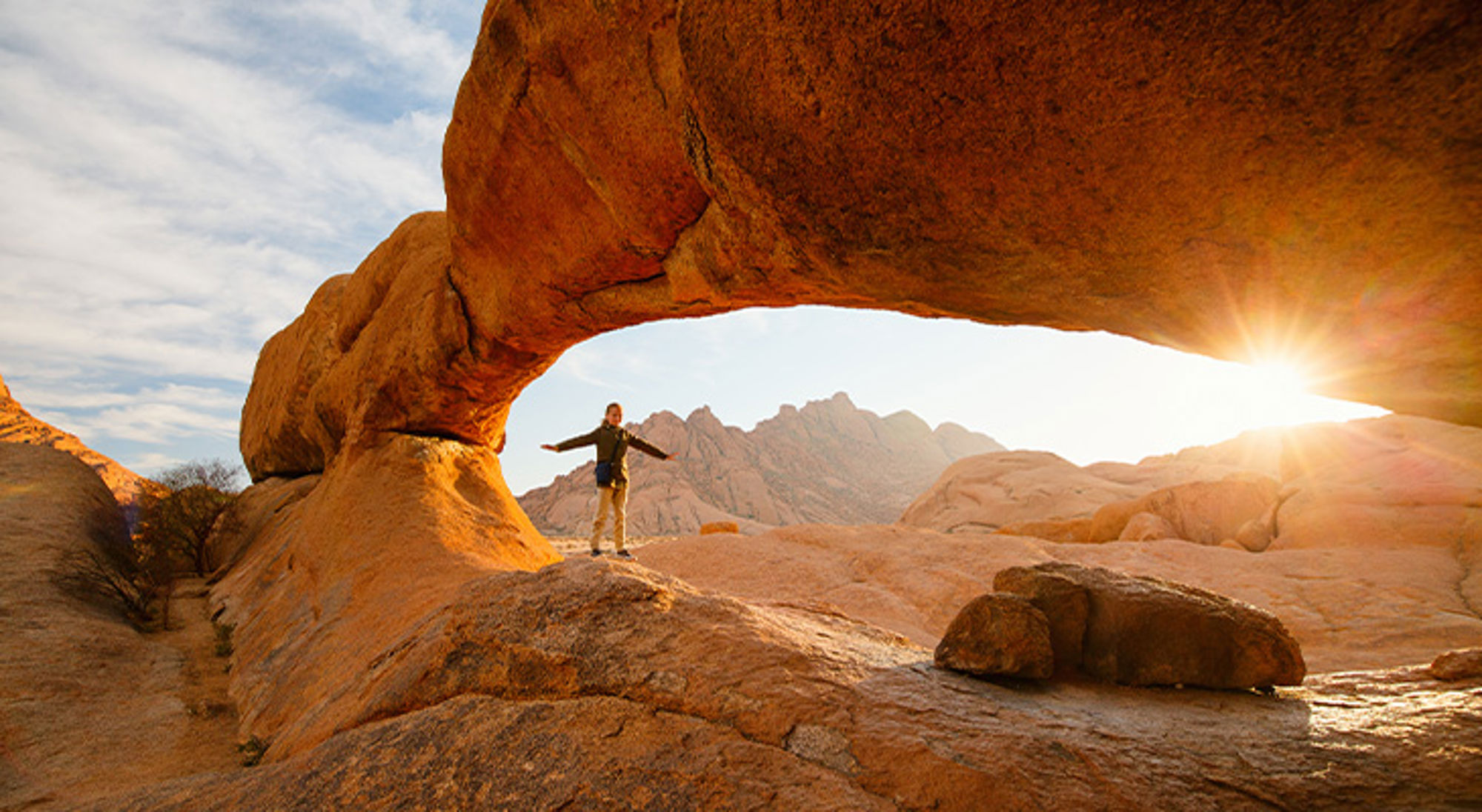 Spitzkoppe Namibië Wandelaar onder rotsboog in Namibië bij zonsondergang, met bergen op de achtergrond.