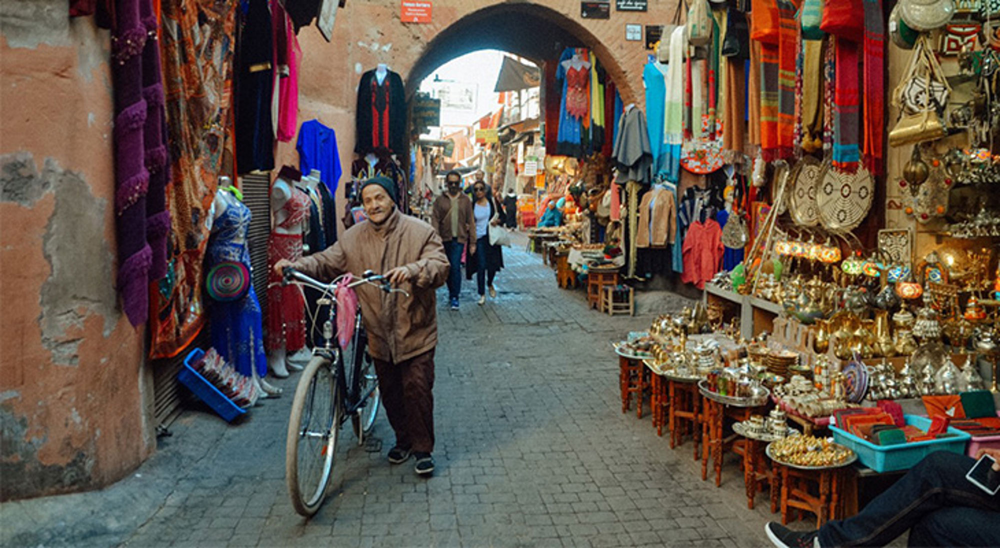 Man met fiets in een smalle souk met kraampjes vol textiel en lampen.