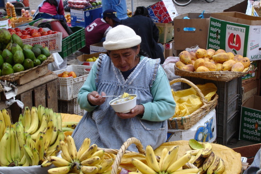 Marktvrouw eet uit een kom tussen stapels bananen, avocado’s en mango’s op een markt in Ecuador.