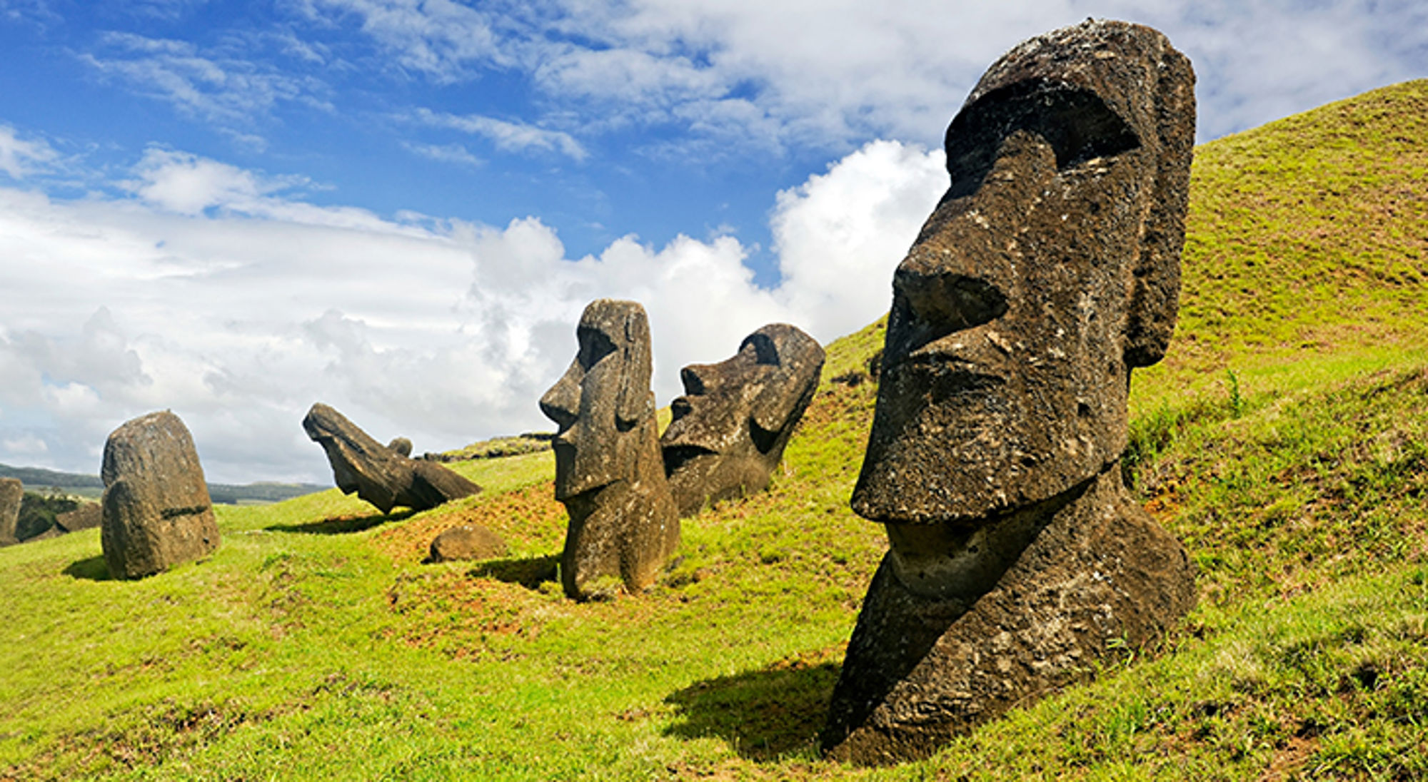 Moai-beelden op Paaseiland op een groene heuvel onder een blauwe lucht met wolken.