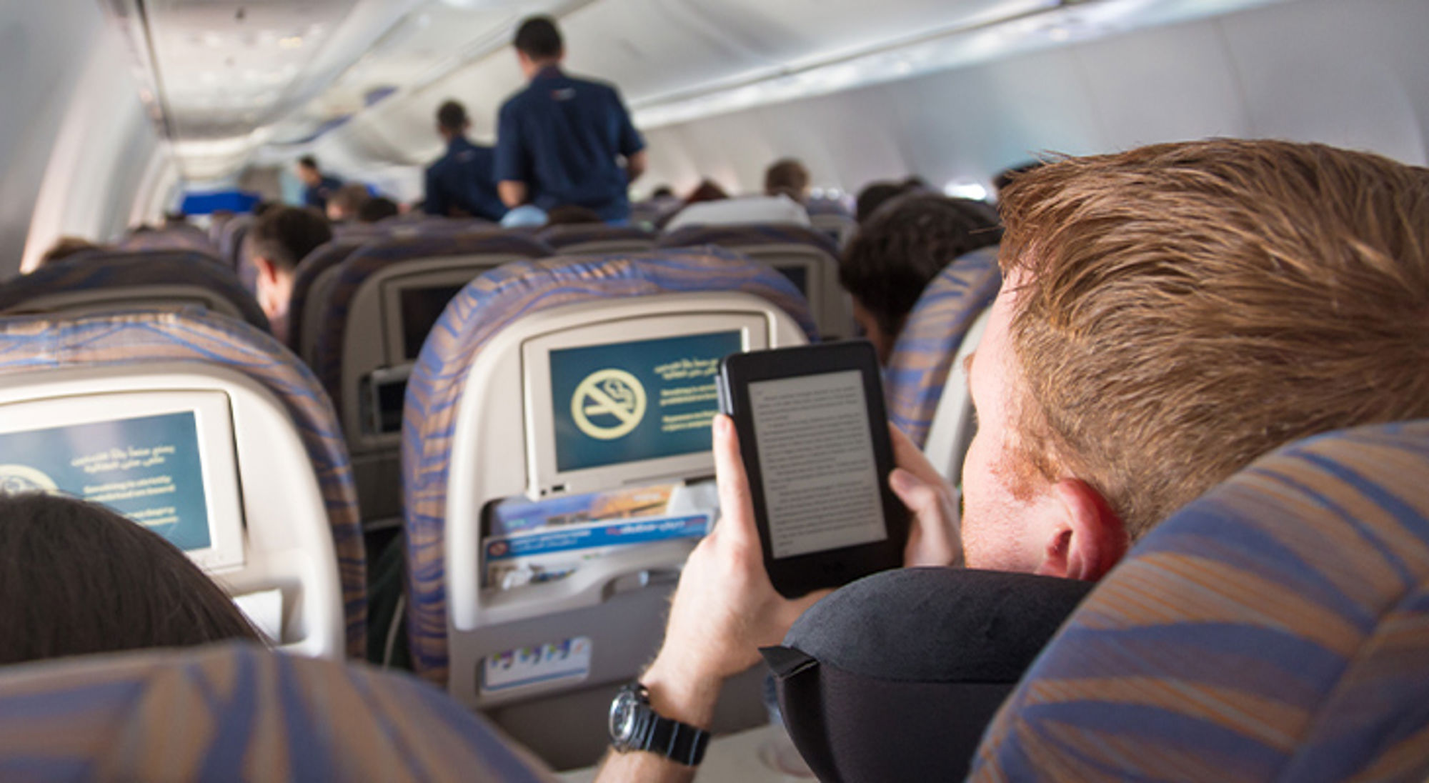 E-reader vliegtuig Passagier leest een e-book op een tablet in het vliegtuig, met rijen stoelen en cabinepersoneel op de achtergrond.