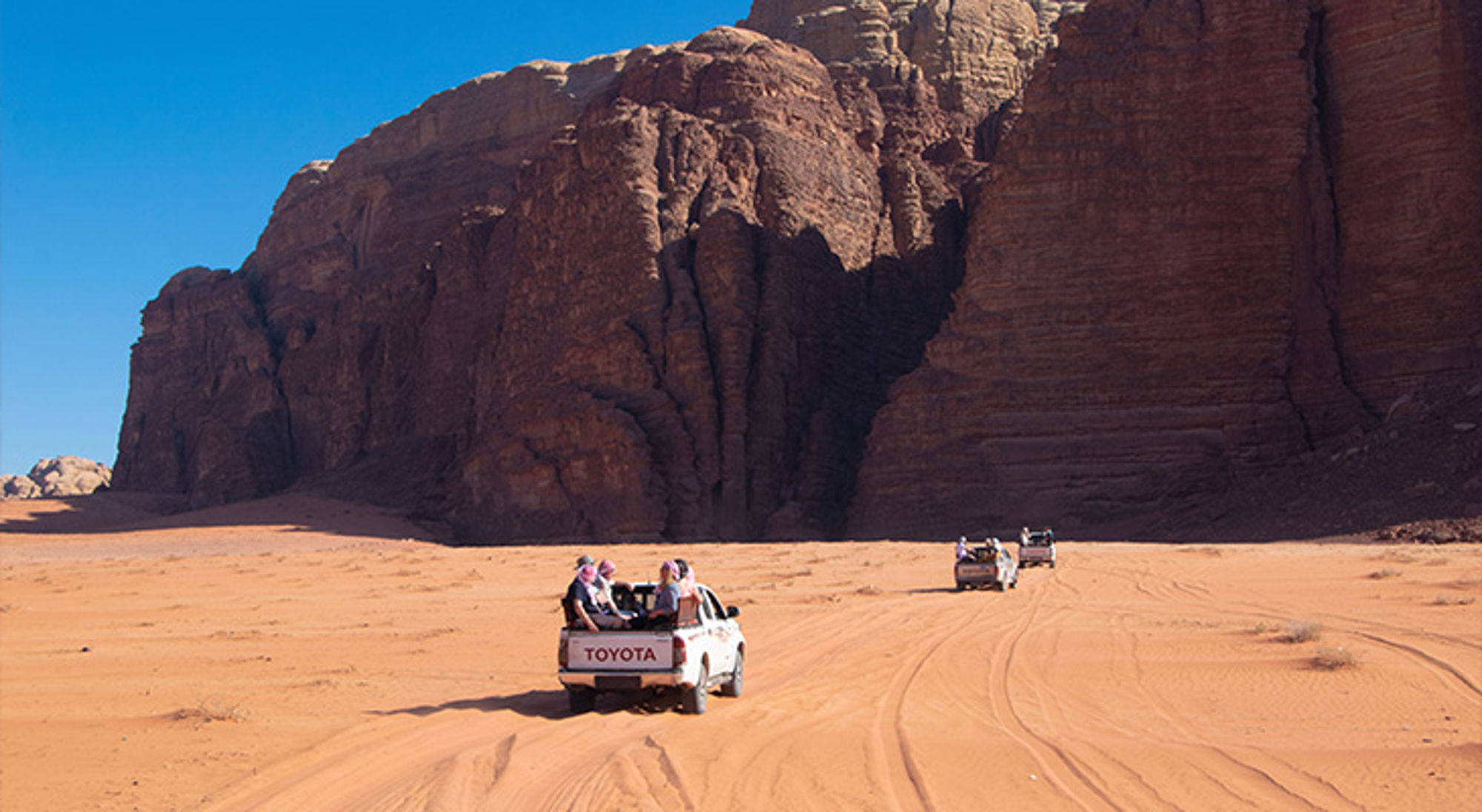 Wadi Rum Jordanië Jeep-safari met pick-uptrucks door de woestijn langs hoge rotsformaties onder een heldere blauwe lucht.