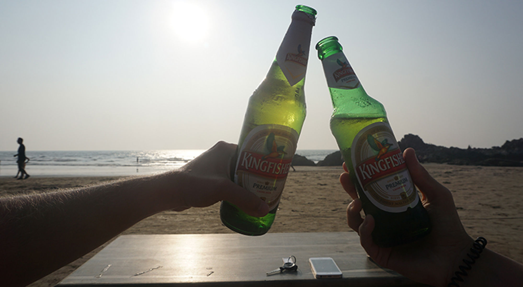 Twee personen proosten met Kingfisher-bier op een strand bij zonsondergang.