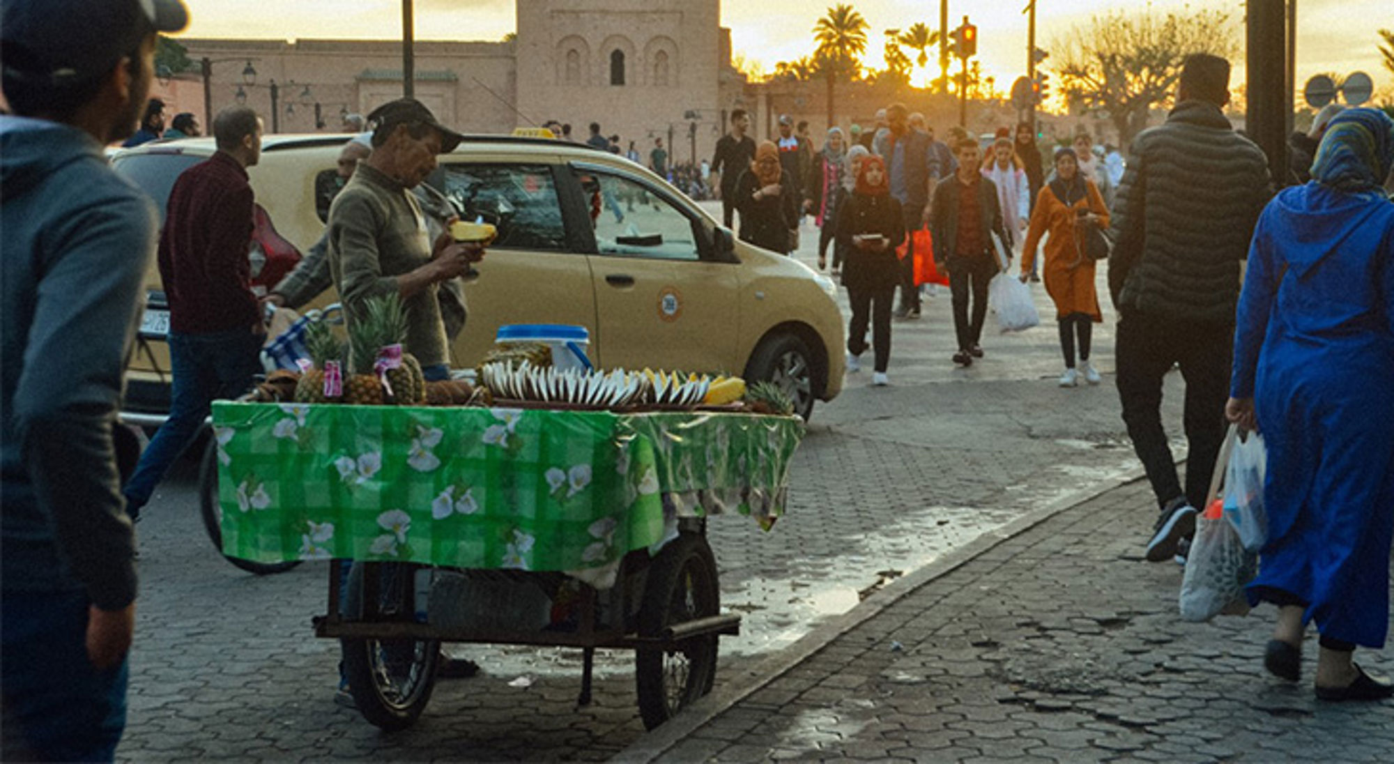 Djemaa El Fna plein met taxi's Straatverkoper met fruitkar en sapjes tussen voorbijgangers op een druk plein bij zonsondergang.
