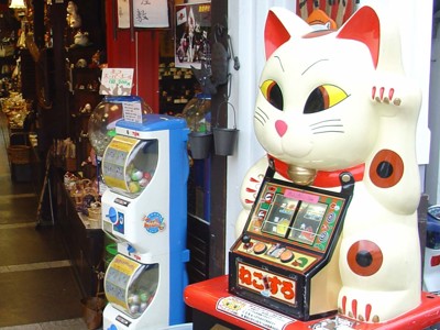 Maneki-neko gelukskat bij een Japanse winkel, naast een gashapon-automaat.