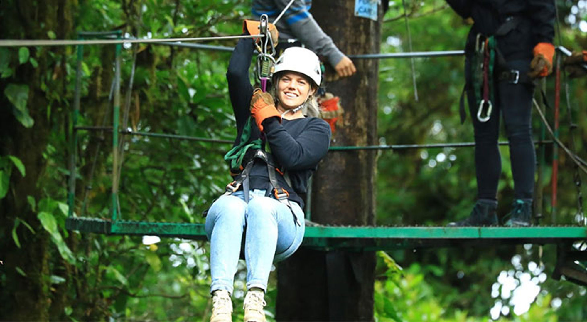 Vrouw met helm en harnas aan een zipline in een groen bos, klaar om te tokkelen.