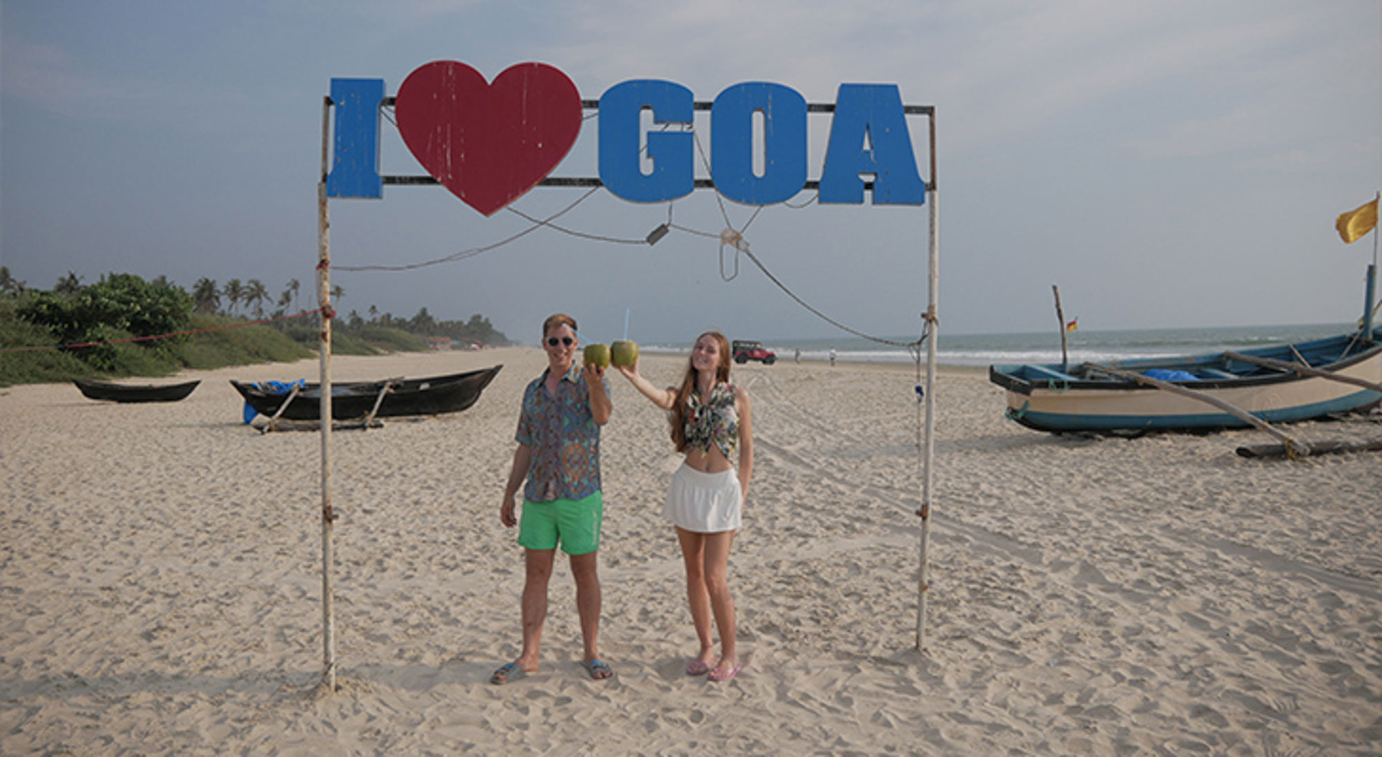 Twee reizigers proosten met kokosnoten op het strand onder het bord “I love Goa”, met vissersboten op de achtergrond.