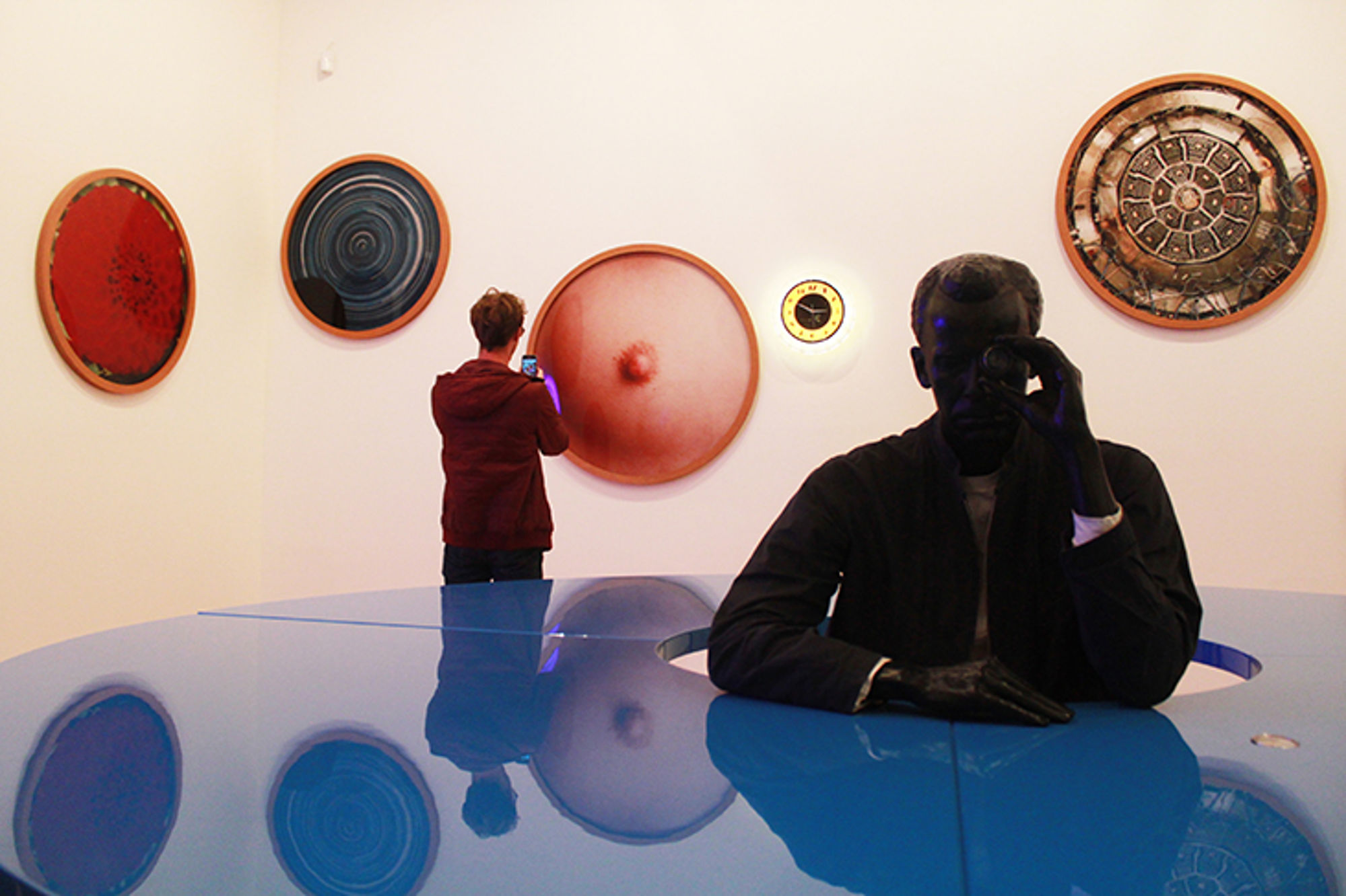 Bezoekers bekijken moderne kunst in een museumzaal met ronde wandobjecten en een blauwe spiegelende installatie.