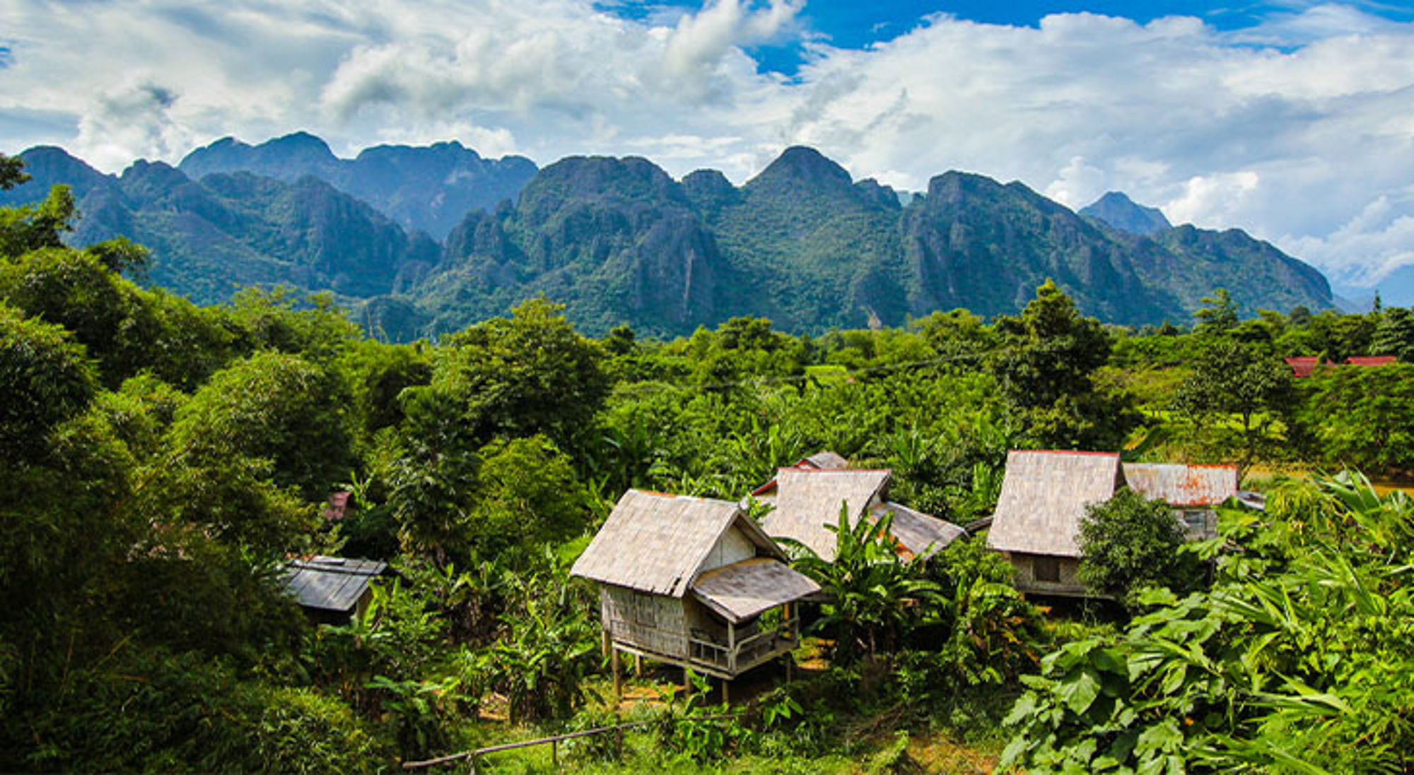 Laos Groen berglandschap met houten huisjes tussen tropische vegetatie onder een blauwe lucht.