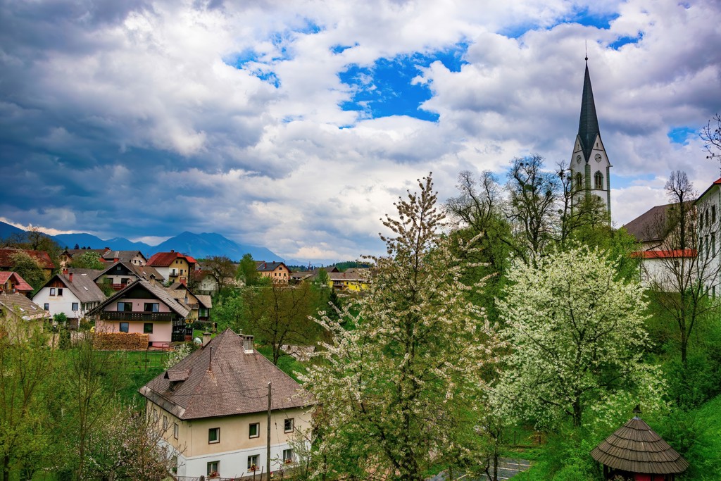 Dorpsgezicht in Slovenië met kerkspits, bloeiende bomen en bergen op de achtergrond.