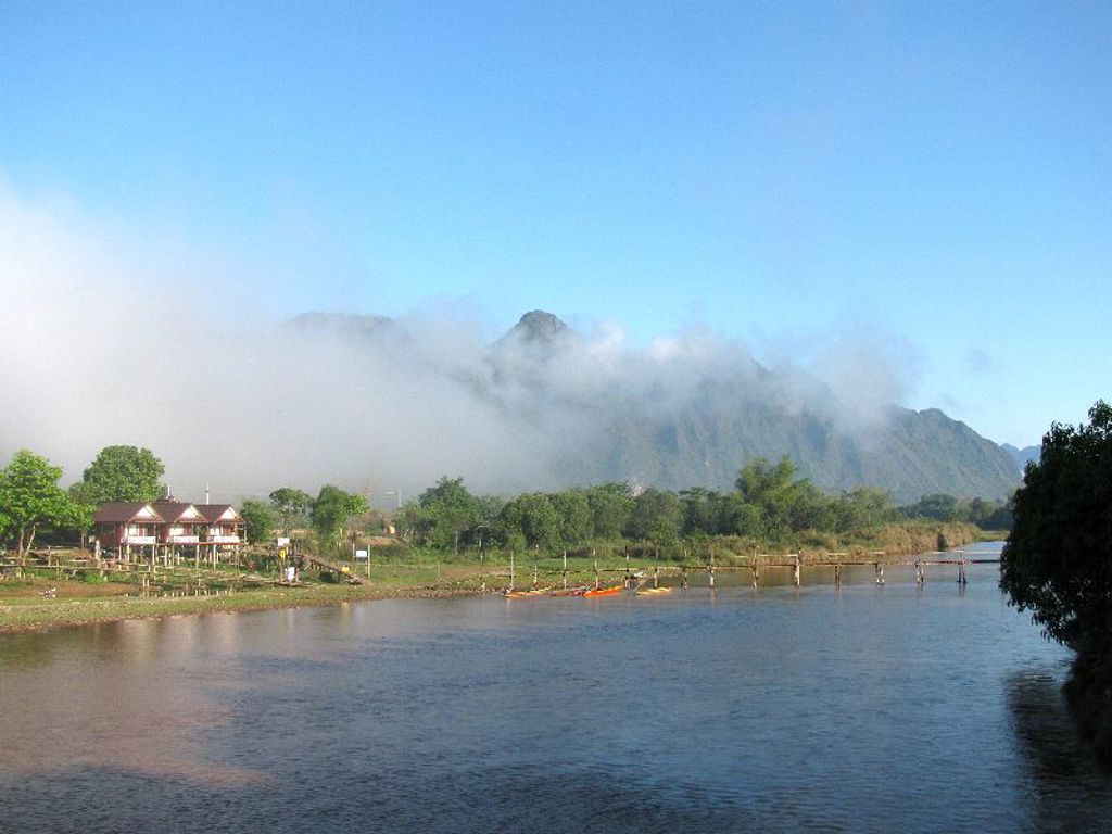 Rivierlandschap in Laos met mistige bergen op de achtergrond, huizen aan de oever en een houten brug.