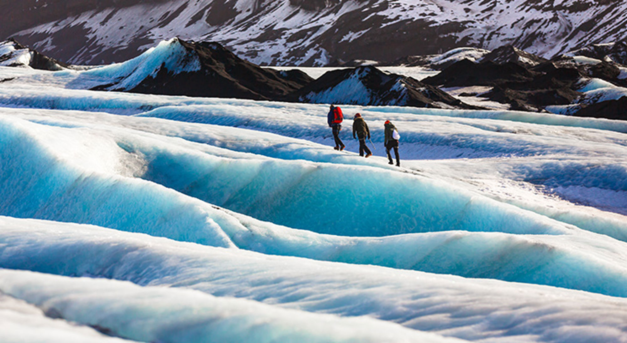Gletsjerwandeling in IJsland Wandelaars trekken over een blauwe gletsjer met besneeuwde bergen op de achtergrond.