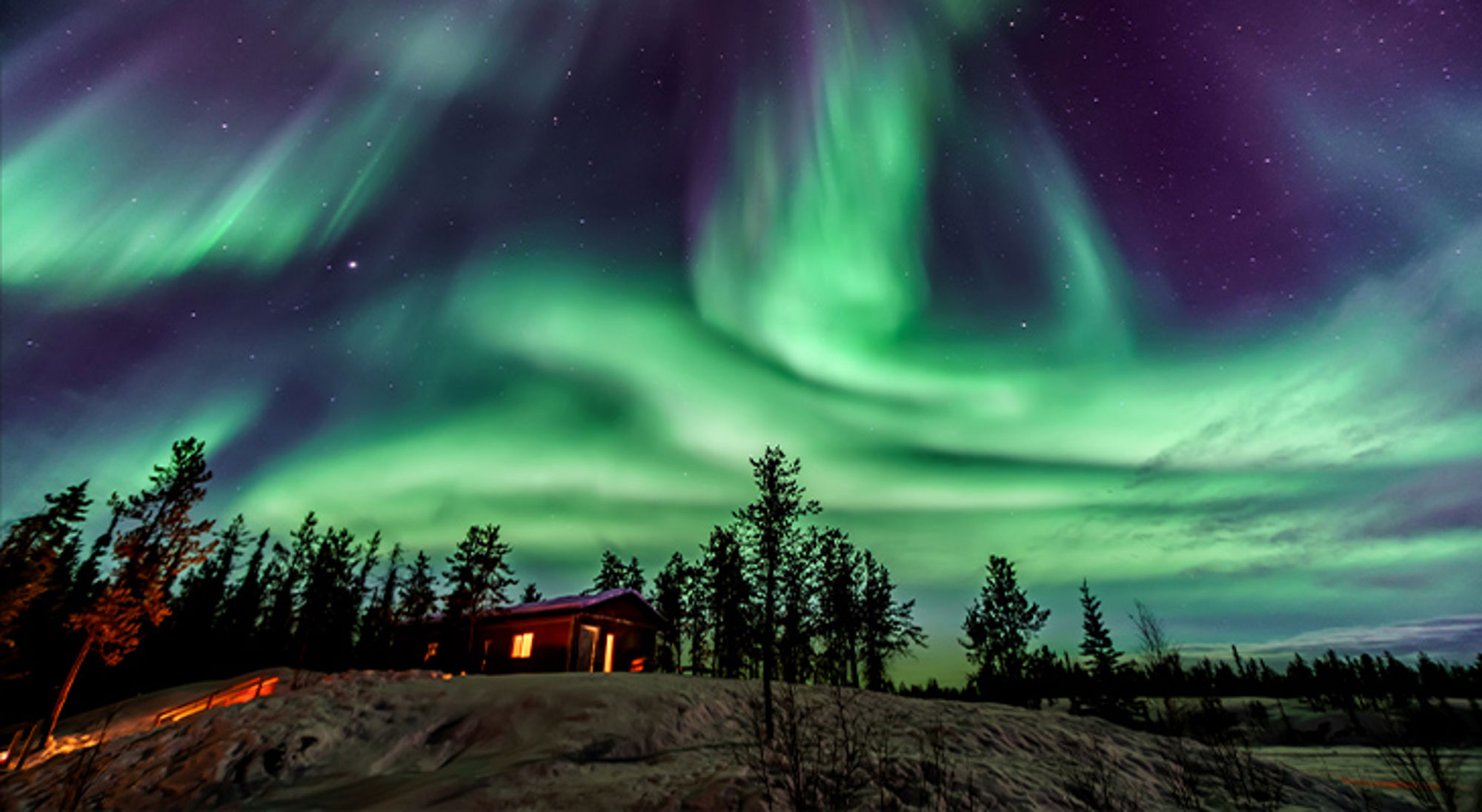 Noorderlicht in Yellowknife, Canada Noorderlicht boven een besneeuwd landschap met dennenbomen en een verlichte houten hut.