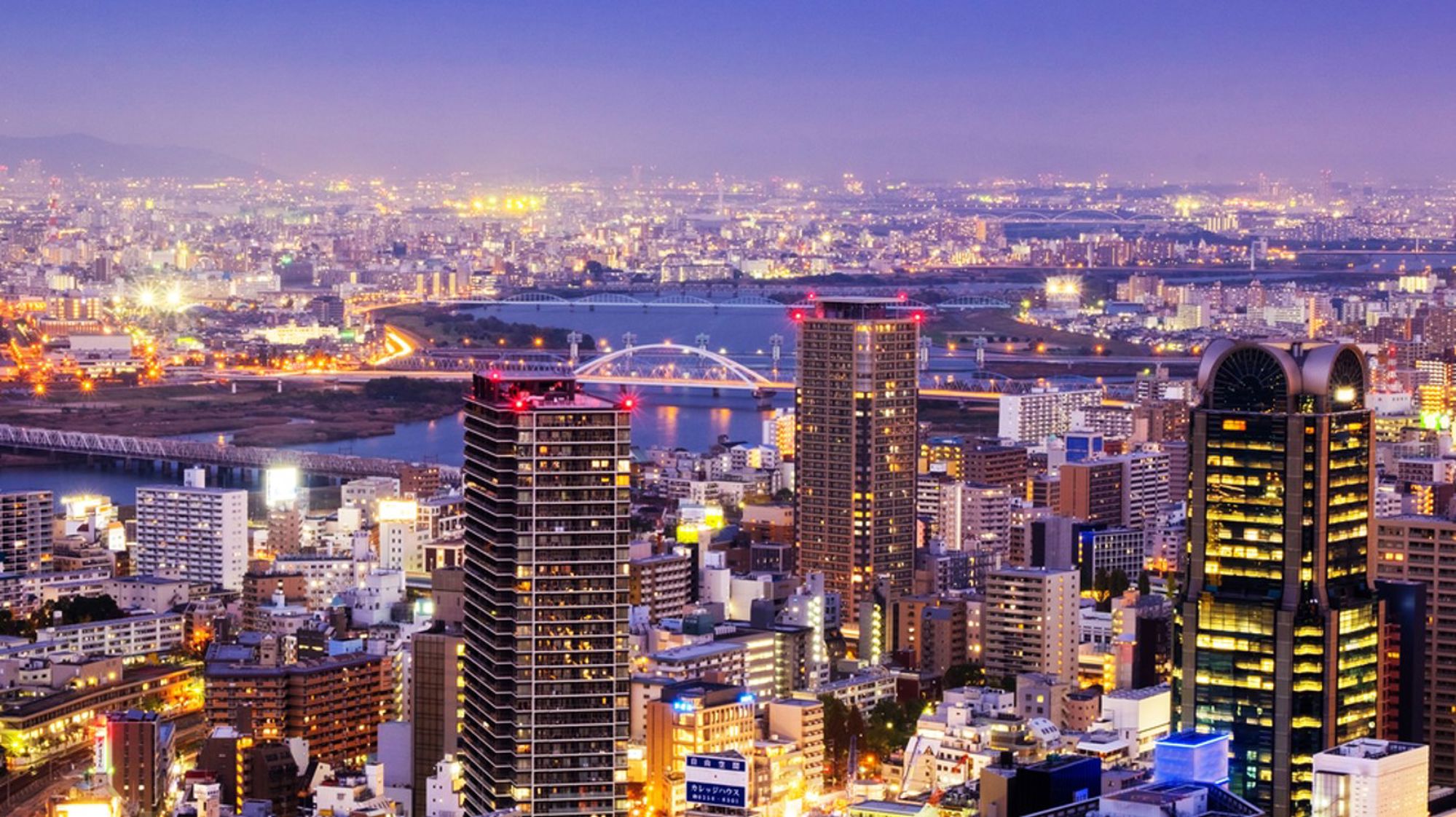 Avondzicht op de skyline van Osaka met verlichte wolkenkrabbers, rivier en bruggen.