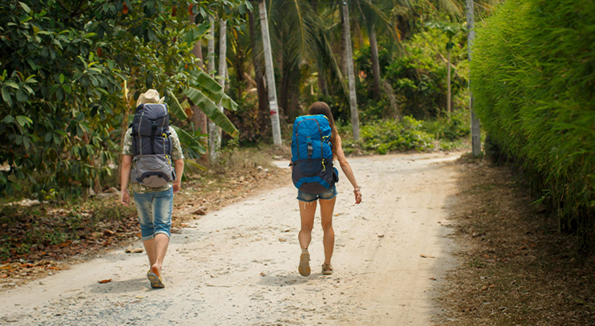 Reizen met een backpack Twee backpackers wandelen over een onverharde weg door tropisch groen.