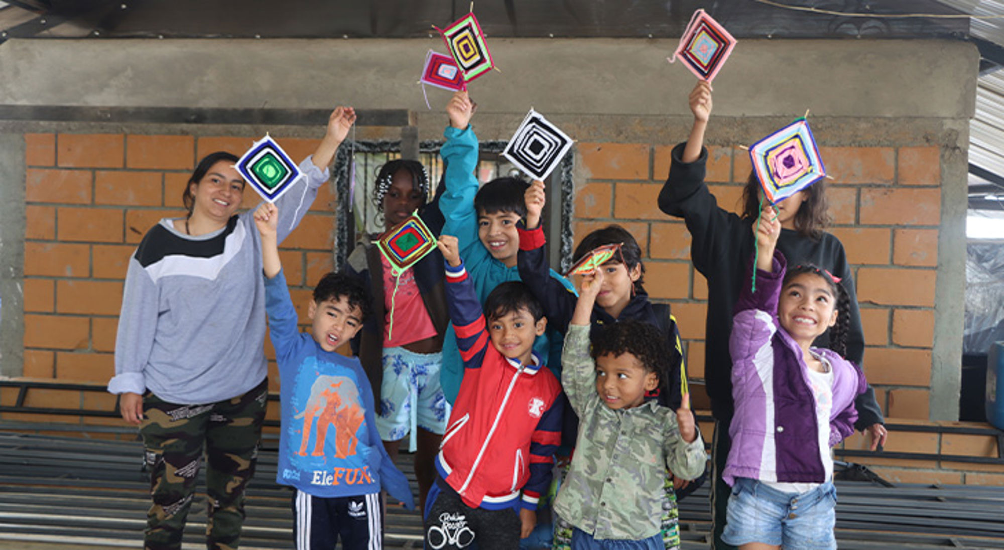 Knutselen Stichting Children of Medellín Kinderen en begeleider houden kleurrijke handgemaakte weefwerkjes omhoog tijdens een creatieve workshop.