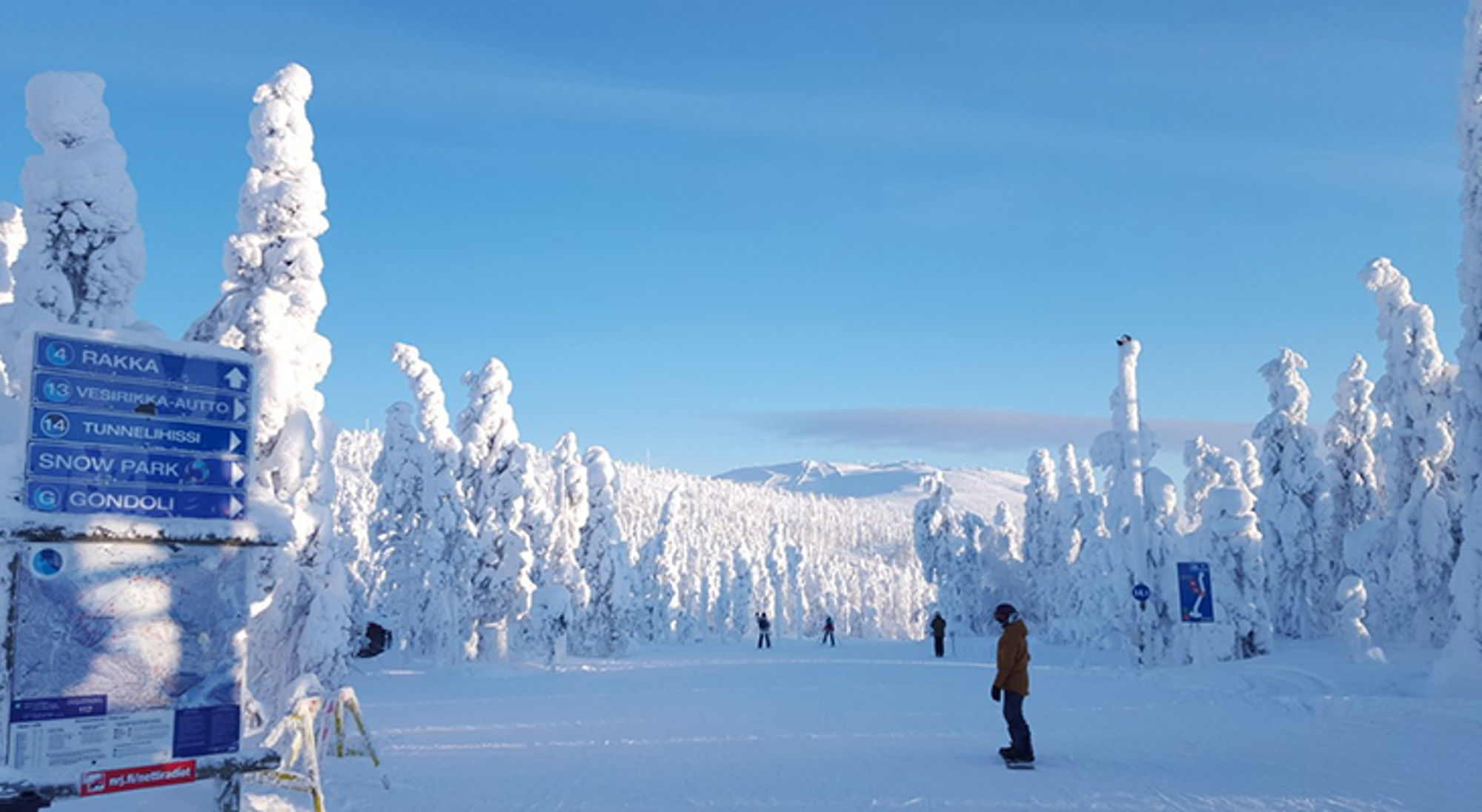 Sneeuwlandschap met besneeuwde bomen, skiërs en een wegwijzer naar Rakka, Snow Park en gondel.