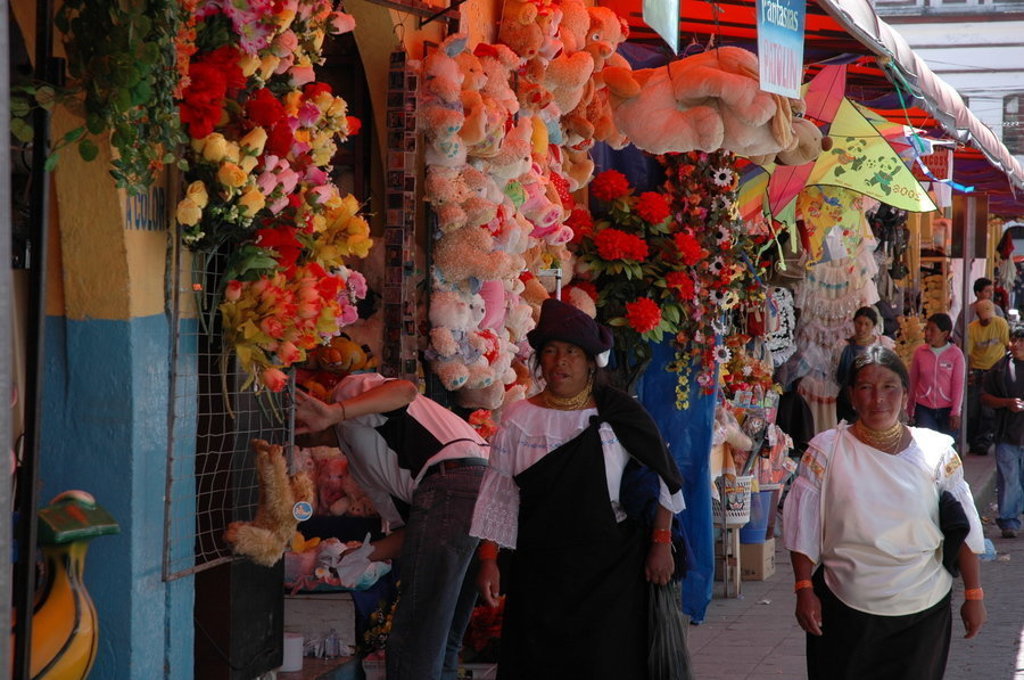 Vrouwen in traditionele kleding op een kleurrijke markt in Ecuador, tussen bloemen, knuffels en kraampjes.