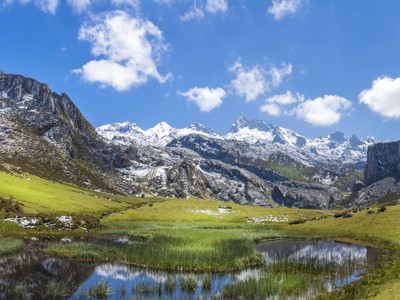 Berglandschap in Noord-Spanje met besneeuwde toppen, groene valleien en een meertje onder een blauwe lucht.