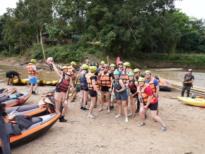 Groep reizigers met zwemvesten en helmen bij opblaasboten aan de rivier tijdens een raftingexcursie in Thailand.