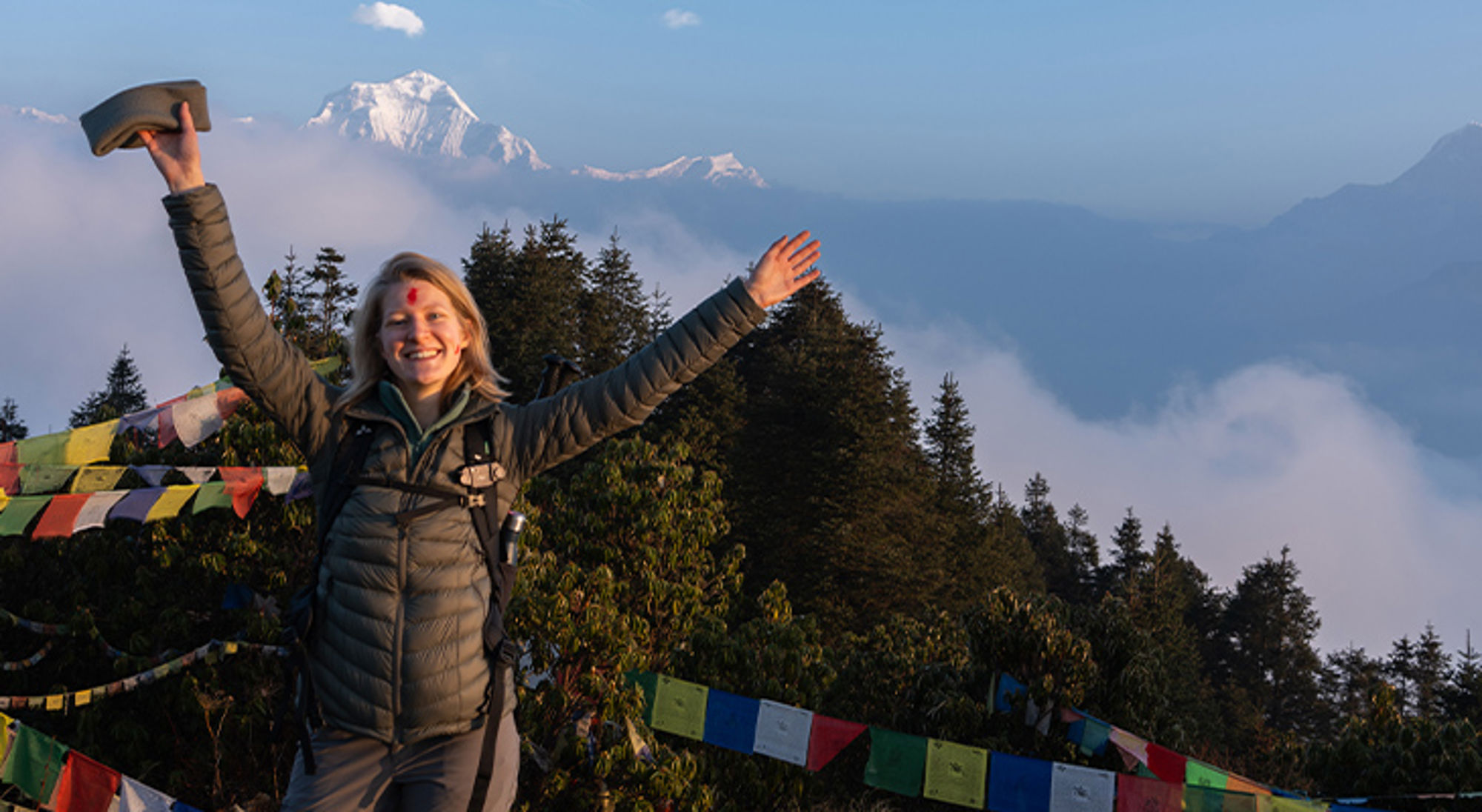 Lisanne Poon Hill Wandelaar met opgeheven armen bij gebedsvlaggen met besneeuwde bergtoppen op de achtergrond.