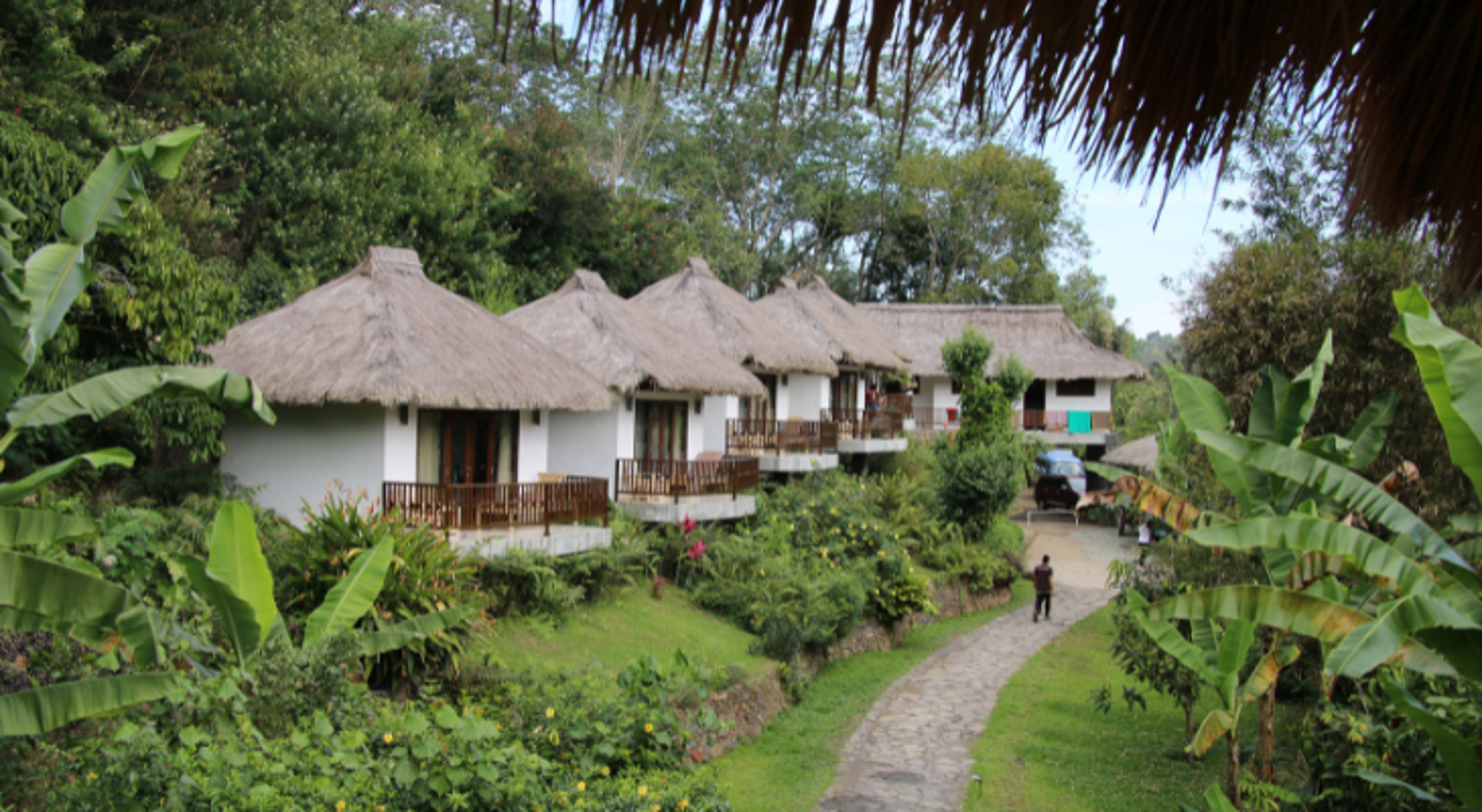 Bungalows met rieten daken en balkons in een groene tropische tuin langs een wandelpad.