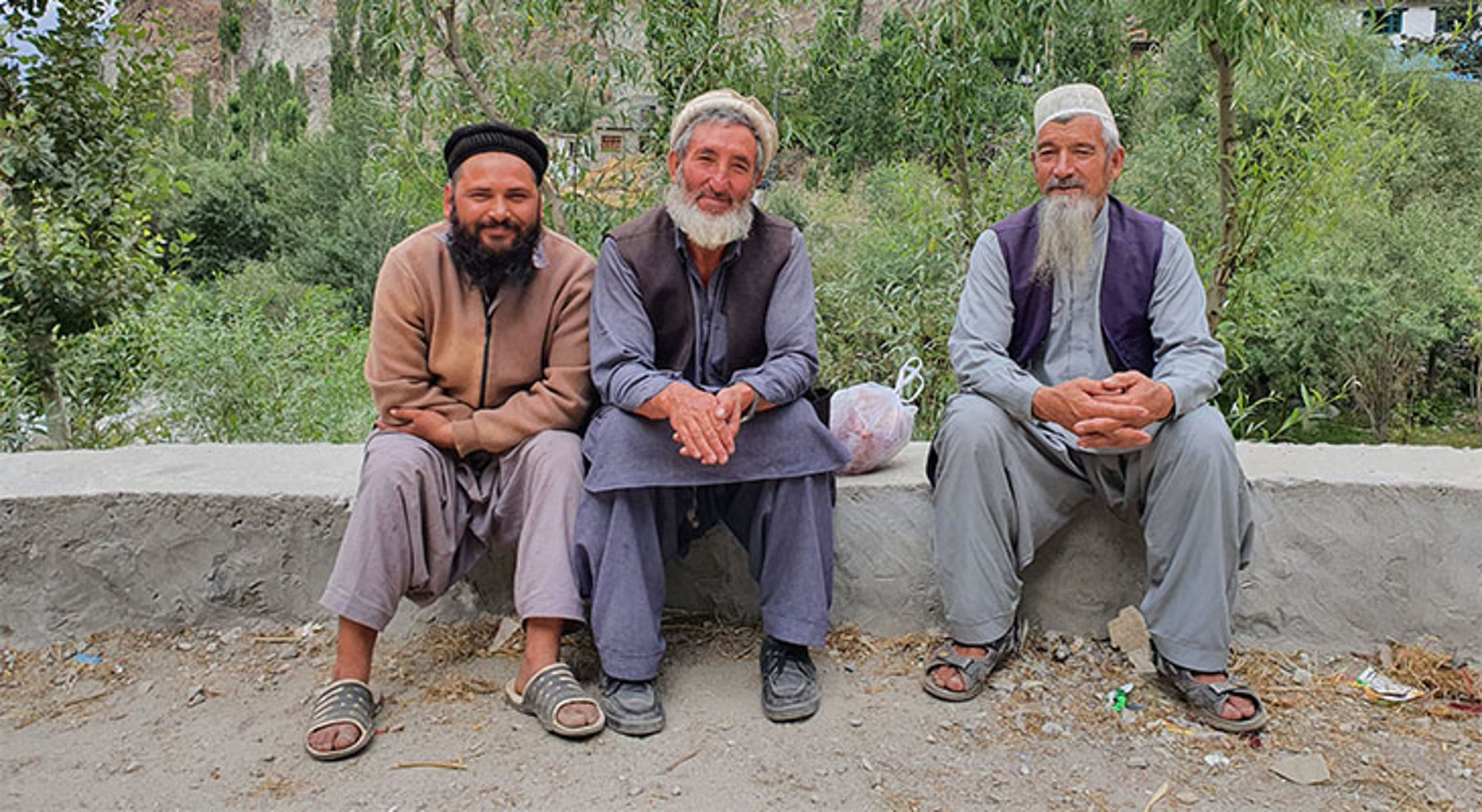 Hunza Vallei Pakistan Drie mannen zitten op een muurtje langs een groene vallei met bergen op de achtergrond.