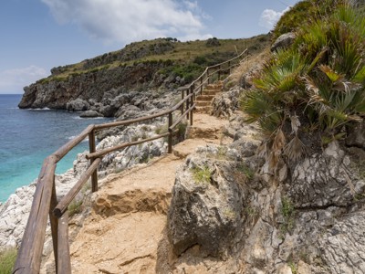 Kustwandeling langs rotsen en helderblauwe zee in West-Sicilië, met pad en houten railing.