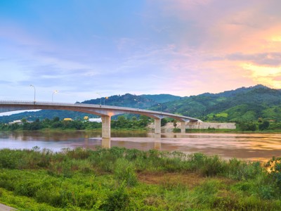 Brug over de Mekong bij zonsondergang, met groene heuvels op de achtergrond in Laos.
