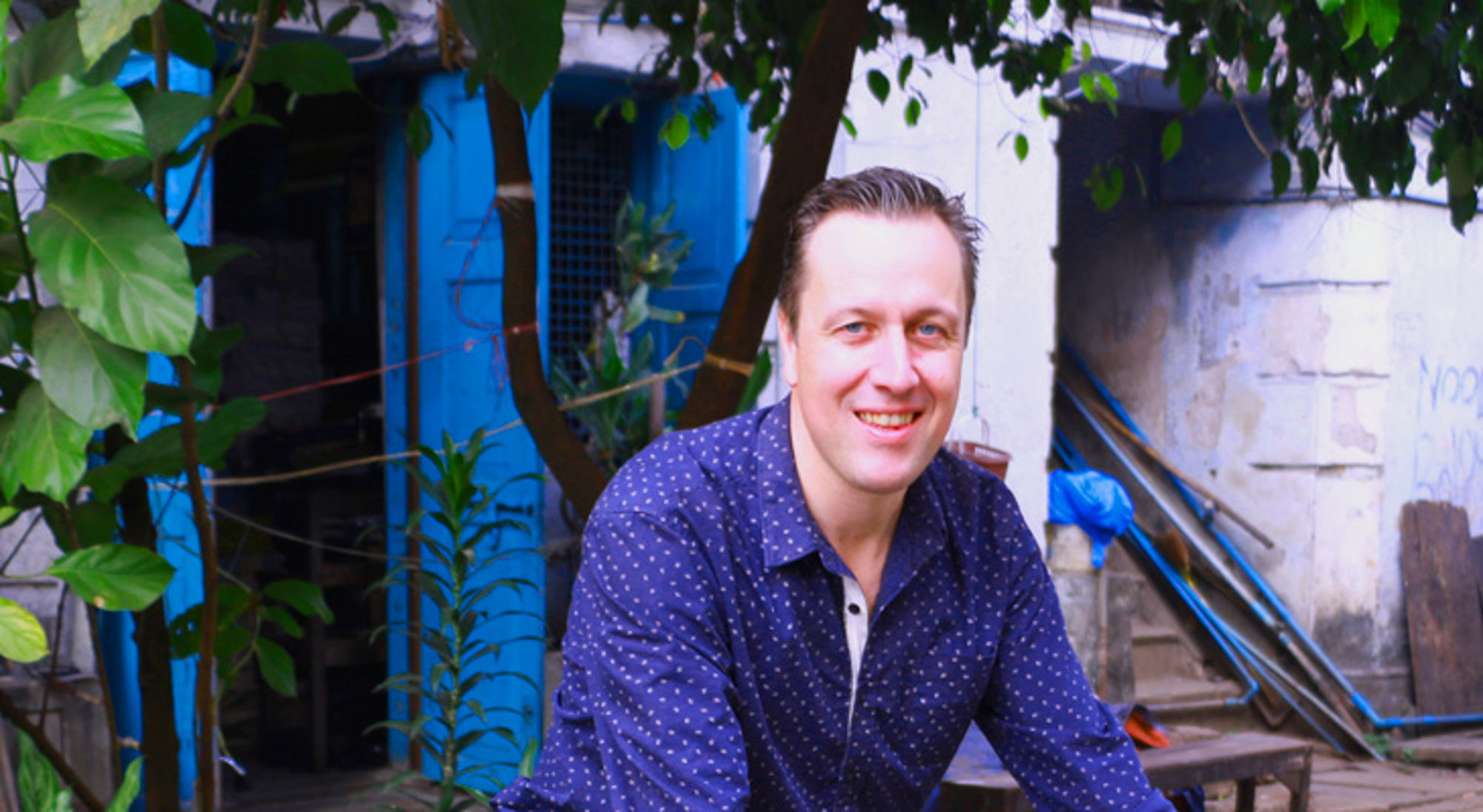 Edwin in Myanmar Portret van een glimlachende man in een blauwe blouse in een tuin met groene bladeren en blauwe achtergrond.