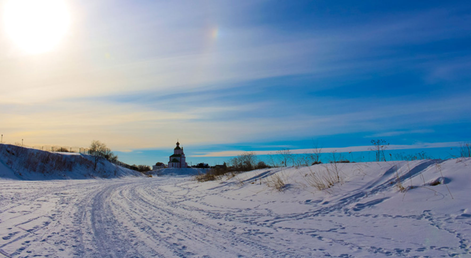 winterlandschap Rusland Besneeuwd landschap met kronkelend pad naar een kerk in de verte onder een blauwe winterlucht.