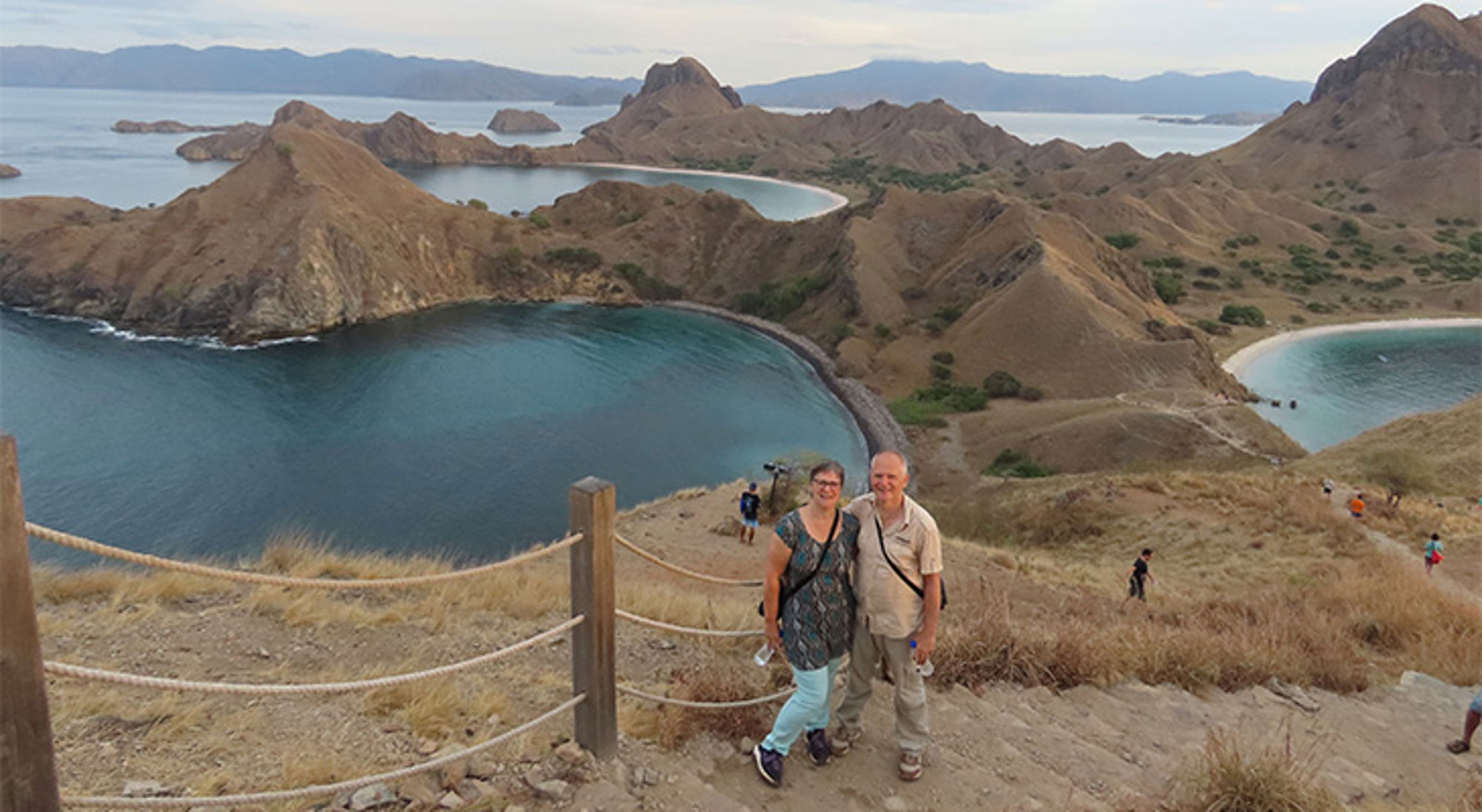 Uitzicht over Komodo National Park met turquoise baaien en heuvels, met twee reizigers op het uitkijkpunt.