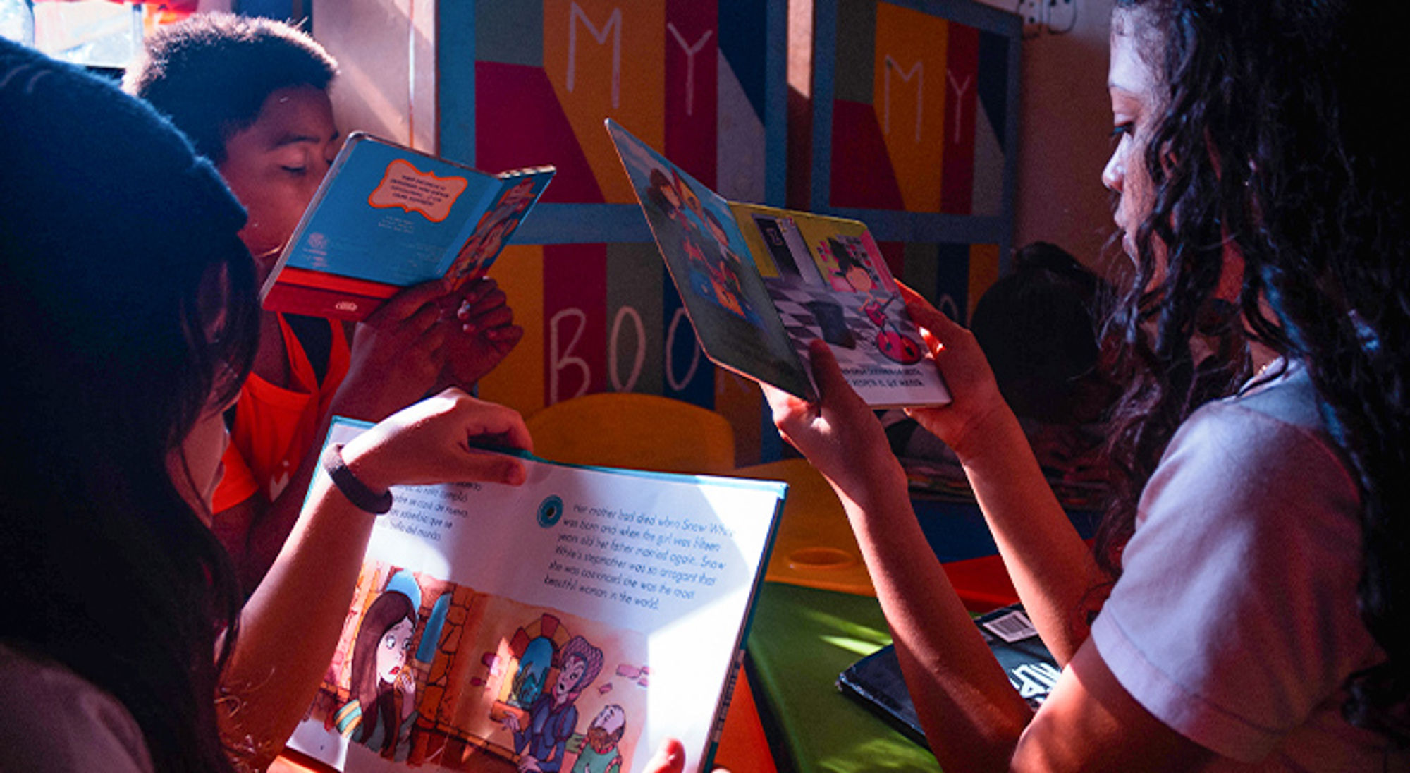 Engelse les Stichting Children of Medellín Kinderen lezen kleurrijke boeken in een bibliotheekhoek met zonlicht en een muurschildering op de achtergrond.