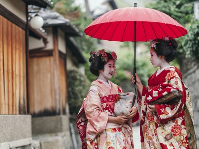 Twee geisha’s in kleurrijke kimono’s onder een rode paraplu in een straat in Japan.