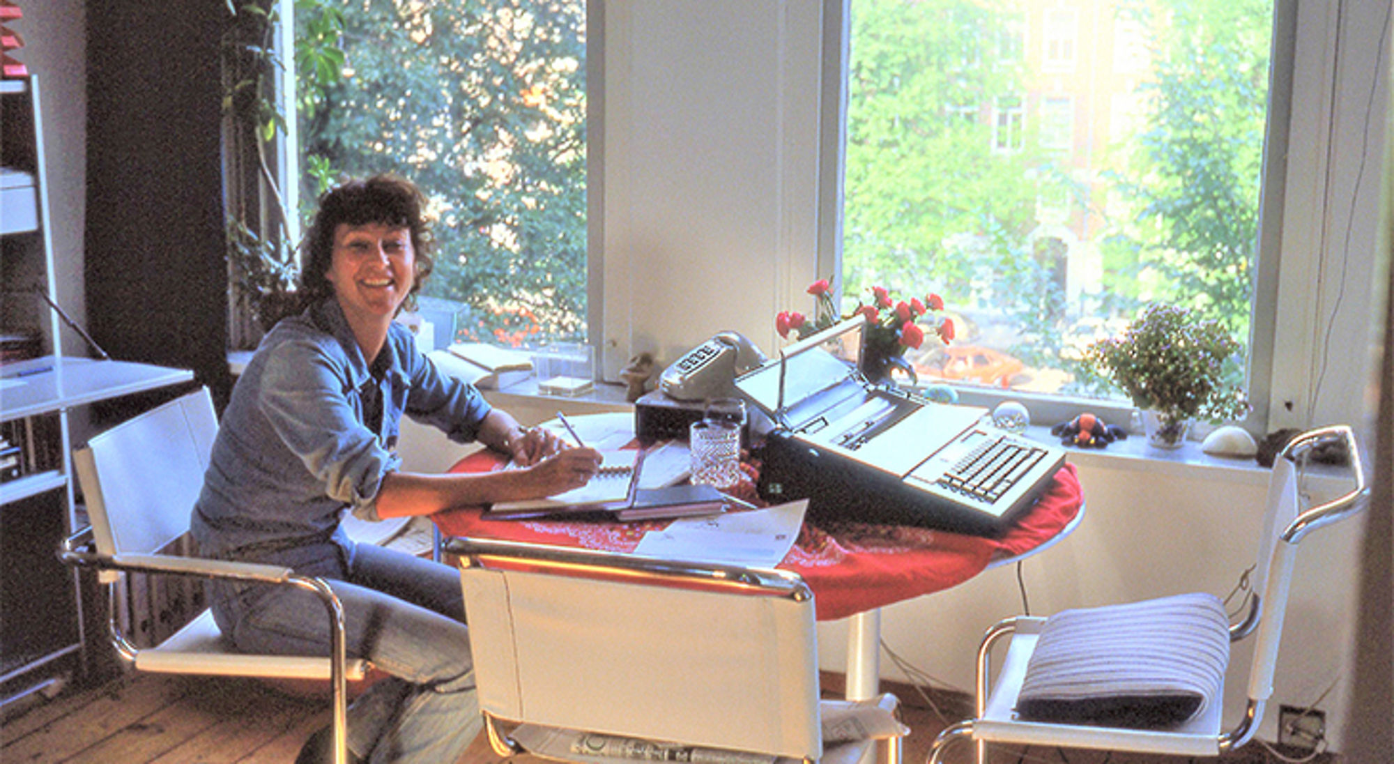 Mieke aan de keukentafel van Ellie Vrouw schrijft aan een tafel bij het raam, met een typemachine en papieren op de achtergrond.