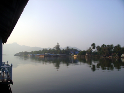 Rustig rivierlandschap met palmbomen en huizen aan de oever, met bergen op de achtergrond in Thailand.