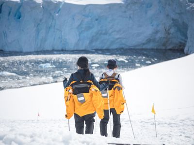 Wandelaars in gele jassen lopen over een sneeuwveld langs een gletsjermeer in Groenland.