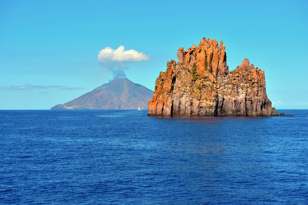 Vulkaan Stromboli met rookpluim achter rotsformatie in zee bij de Eolische Eilanden, Sicilië.