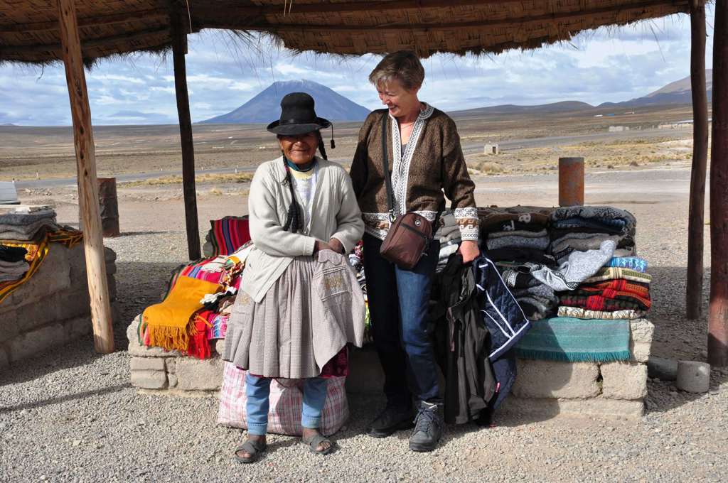 Vrouw op reis naar Peru bij een markt met lokale verkoper en kleurrijke dekens, met bergen op de achtergrond.