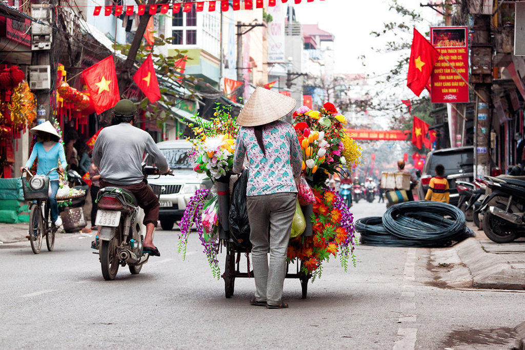 Bloemenverkoper met handkar in een drukke straat in Hanoi, Vietnam, met Vietnamese vlaggen.