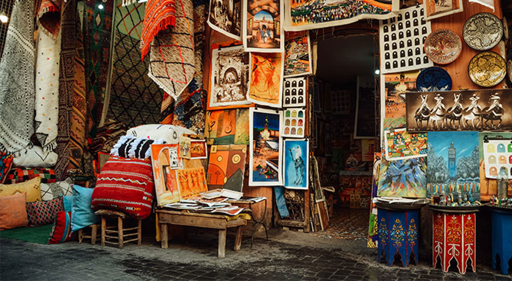 Kunst in Aït-Ben-Haddou Kleurrijke souk met tapijten, kussens en kunstprints bij een winkelingang.