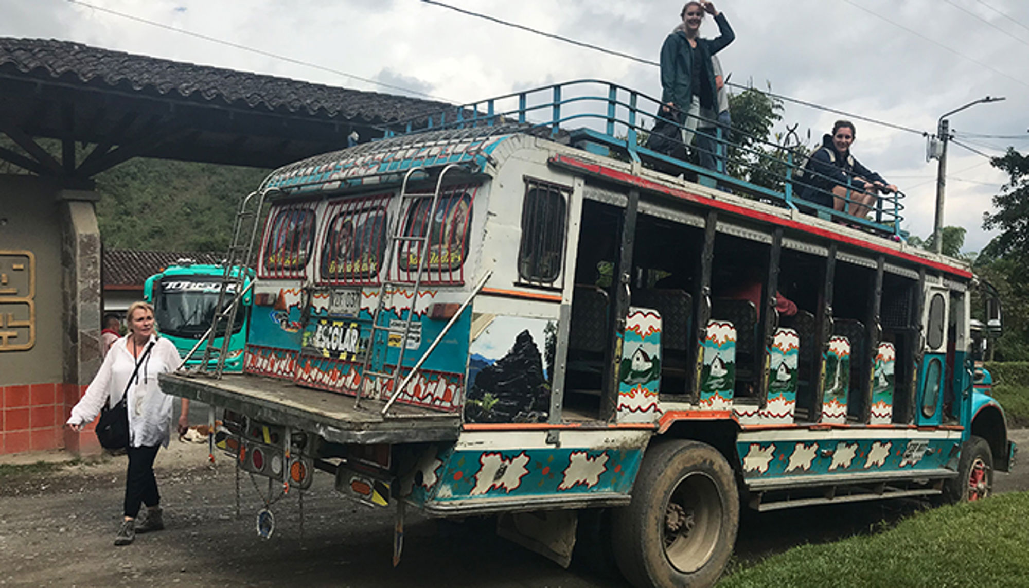 Reizigers op een kleurrijke bus met open zijkant en dakrek bij een halte in de bergen.