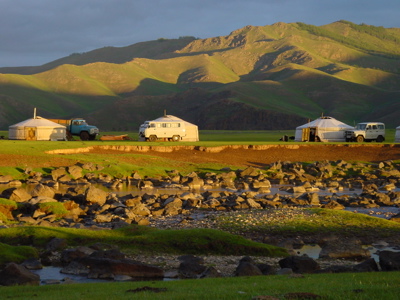 Ger-kamp met ronde tenten en terreinwagens in een groene vallei in Mongolië, bij een rivier en heuvels.