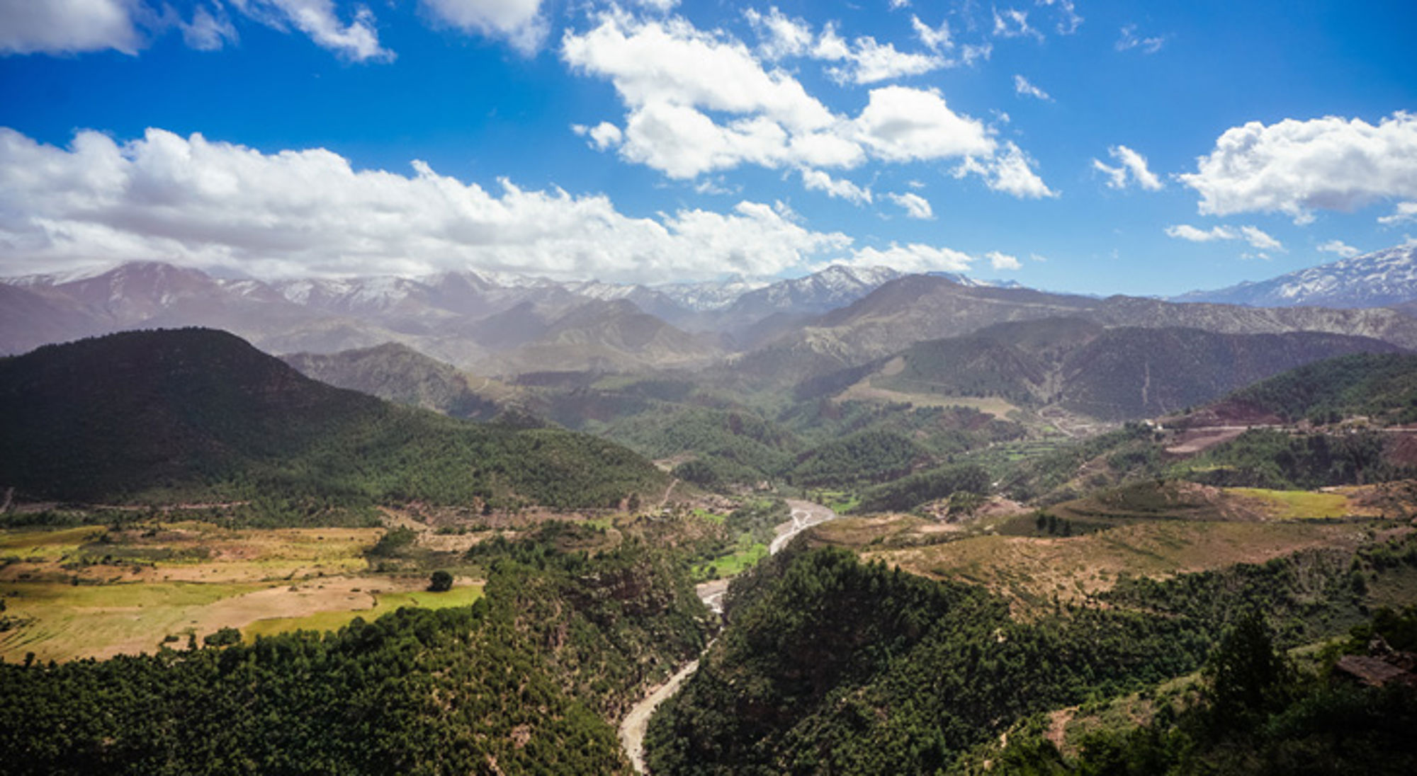 Panoramisch uitzicht op een groene vallei met rivier en bergketen onder een blauwe lucht met wolken.
