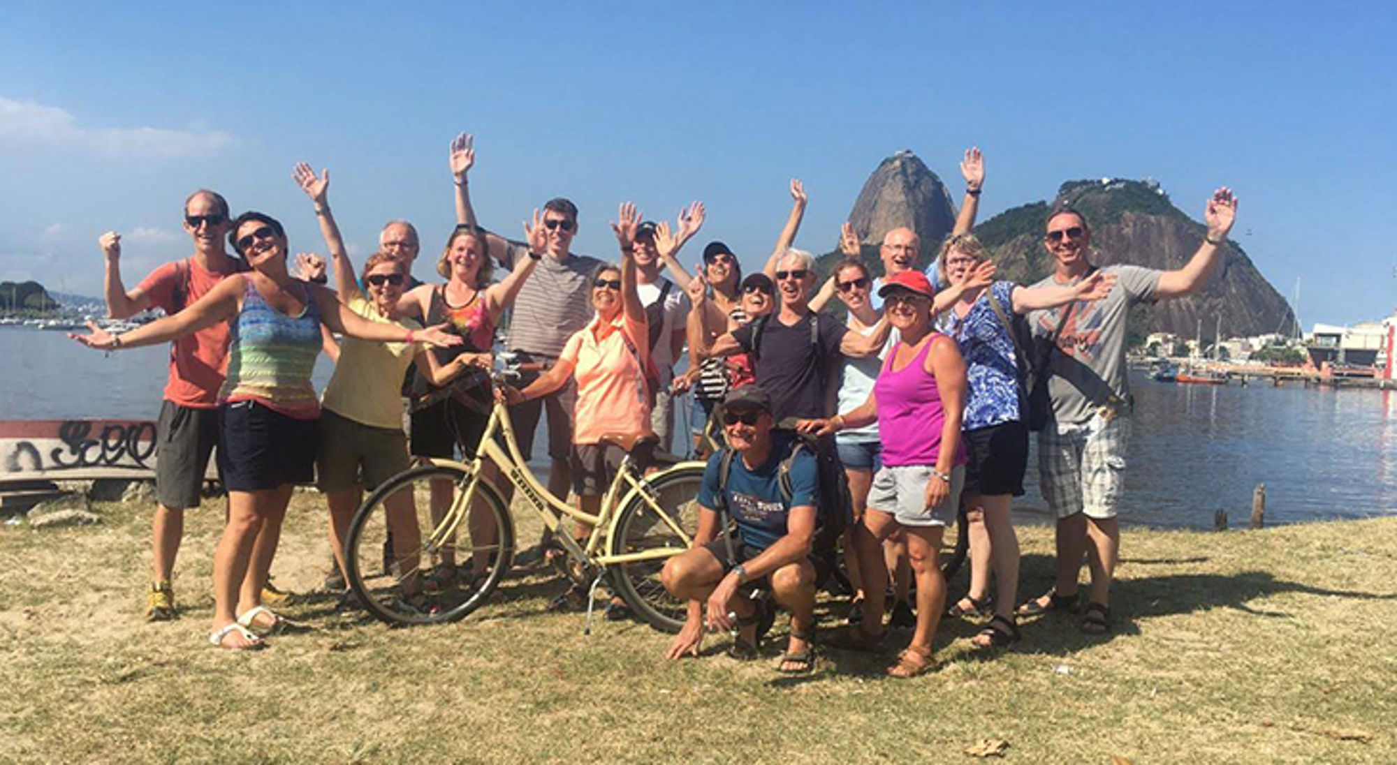 Groep reizigers poseert met fiets aan het water in Rio de Janeiro, met de Suikerbroodberg op de achtergrond.